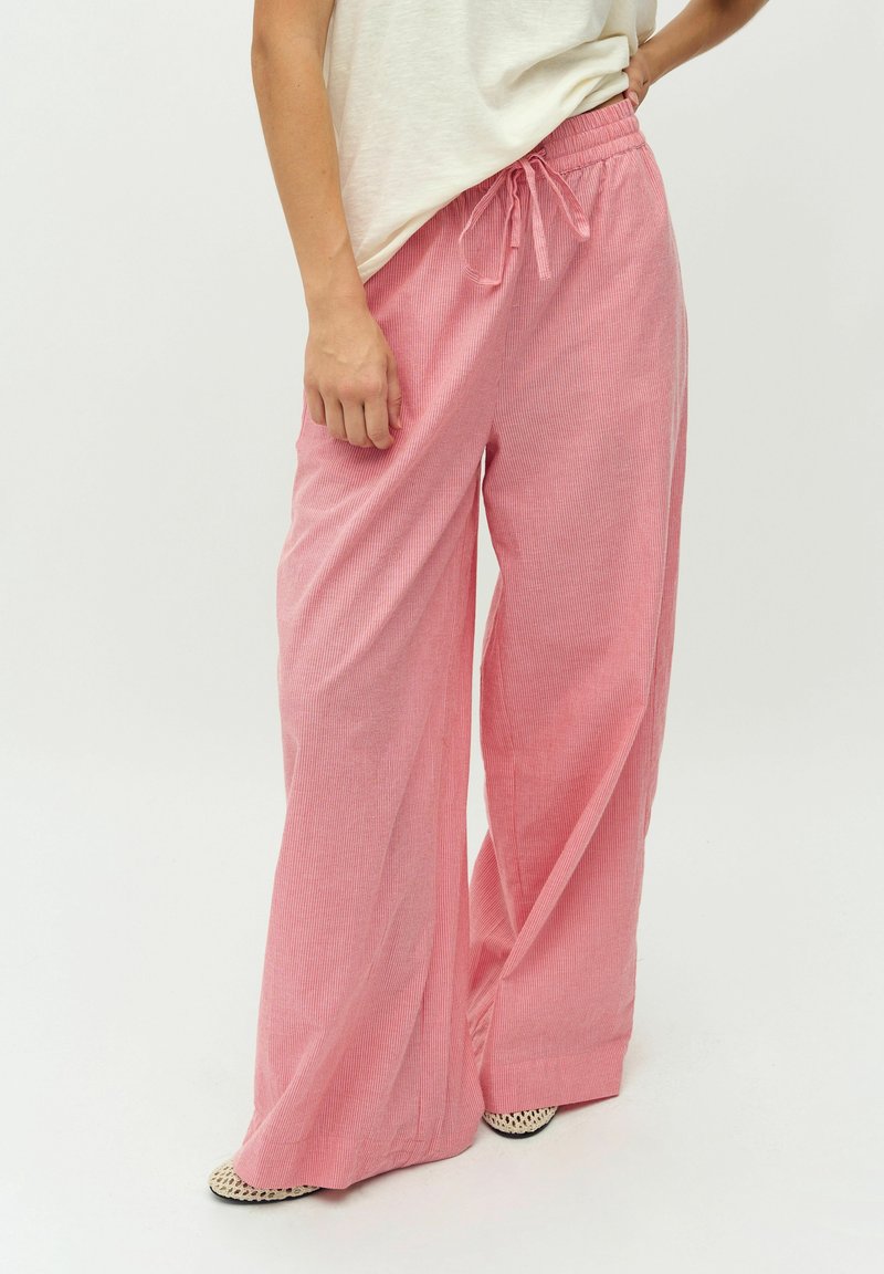 Personne portant un pantalon en velours côtelé rose ample, taille haute avec cordon de serrage à la taille, et un haut blanc, debout avec une main sur la hanche.
