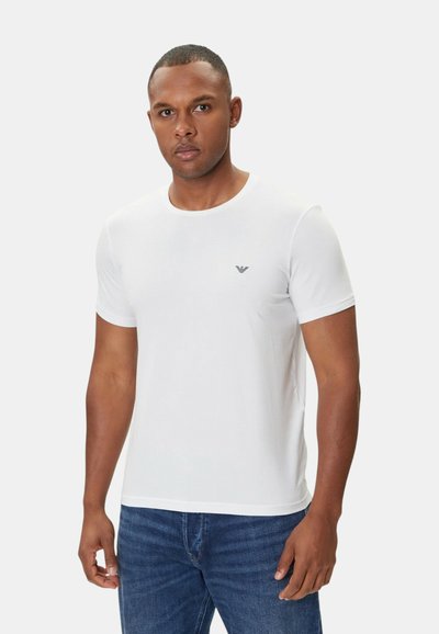 Valkoinen lyhythihainen T-paita pyöreällä kauluksella ja pienellä sinisellä logolla rinnassa, yhdistetty tummansinisiin farkkuihin. Sujuva kangaslaatu.