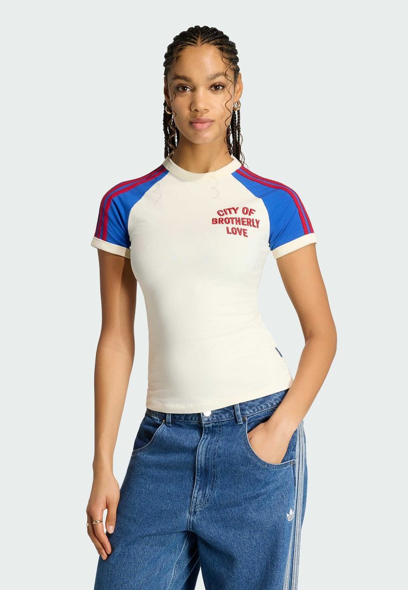 Jeune femme aux cheveux tressés portant un haut à manches courtes blanc et bleu avec l'inscription "City of Brotherly Love" et un jean Adidas bleu.