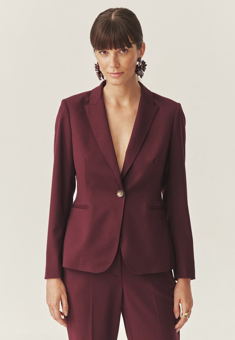Vrouw met donker haar en een pony draagt een bordeauxkleurige blazer op maat en bijpassende broek met grote bloemenoorbellen en een gouden ring.