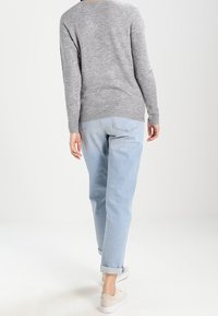 Personne marchant de dos, portant un pull gris à manches longues, un jean bleu clair avec revers et des baskets beiges sur un fond uni.