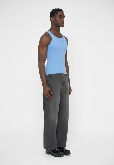 Svetlomodrý nátelník s okrúhlym výstrihom, doplnený o nadmerné sivé džínsy a čierne topánky na hrubom opätku, stojaci na bielom pozadí.