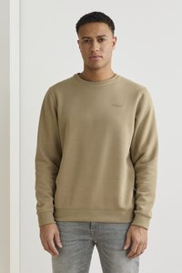 Sudadera beige de cuello redondo con puños y dobladillo de canalé, que presenta un pequeño logotipo bordado en el pecho, combinada con unos vaqueros de color gris claro.