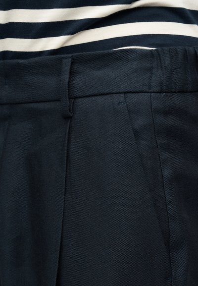 Κοντινό πλάνο σκούρου ναυτικού υφασμάτινου παντελονιού με θηλιές για ζώνη, εμπρόσθιες πιέτες, και ένα λευκό πουκάμισο με ναυτικές ρίγες που είναι μπηγμένο μέσα.