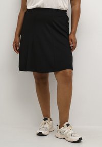 Jupe noire, tissu lisse, forme évasée, longueur genou, avec plis à l'avant. Associée à des baskets blanches et grises avec des accents texturés.