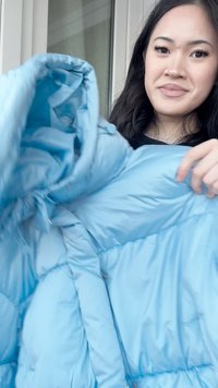 Nuori nainen pitää kädessään ja esittelee vaaleansinistä hupullista toppatakkia sisätiloissa ikkunan lähellä.