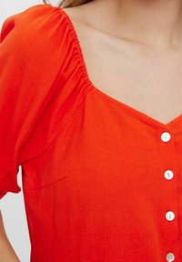 Haut rouge à boutons avec un décolleté carré, manches courtes bouffantes, tissu texturé et petite poche latérale. Présente des boutons blancs et des détails froncés.