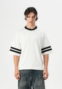 Hvid T-shirt med korte ærmer, sort ribbet rund halslinning og sorte dobbeltstriber på ærmerne. Stoffet ser blødt og tekstureret ud.