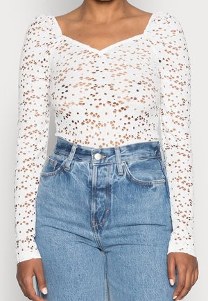 Femme portant un haut en dentelle blanc à manches longues avec des découpes florales et un jean en denim bleu taille haute, les mains détendues le long du corps.