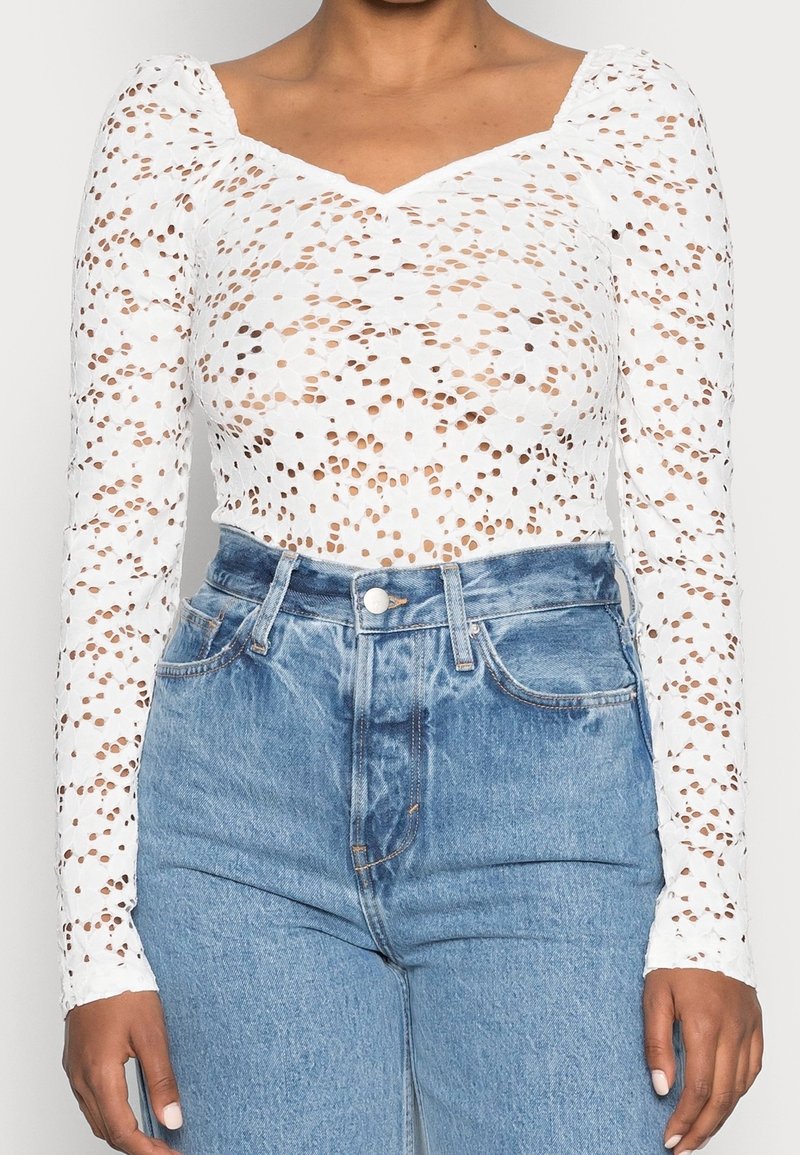 Femme portant un haut en dentelle blanc à manches longues avec des découpes florales et un jean en denim bleu taille haute, les mains détendues le long du corps.