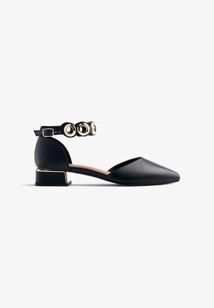 Chaussure plate en simili cuir noir avec un bout pointu, une bride de cheville ornée d'accents circulaires et un talon bloc en métal doré.