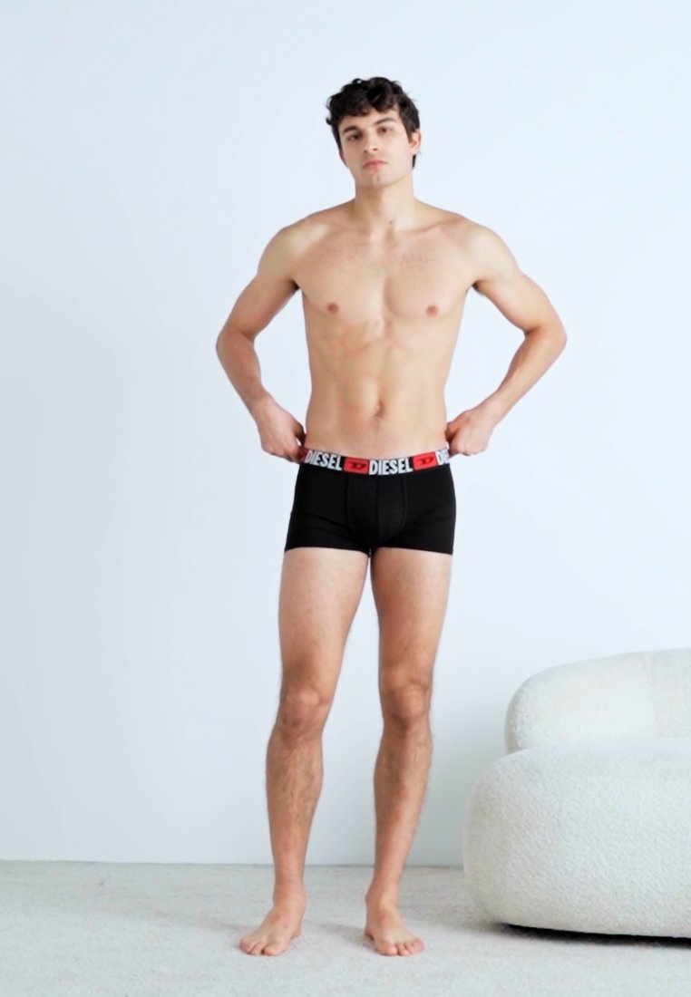 Boxers pour hommes noirs avec un élastique rouge et blanc, arborant le logo "DIESEL" ; modèle debout, mains ajustant l'élastique, pieds nus.