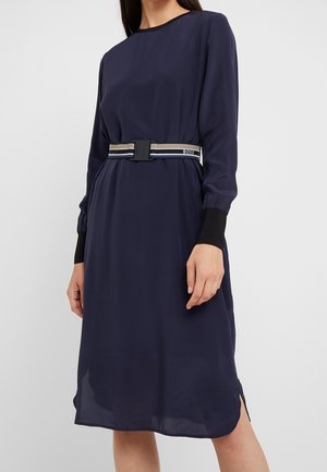 Woman wearing a navy blue long-sleeve dress with black cuff details and a striped belt with a black clasp labeled "BOSS."