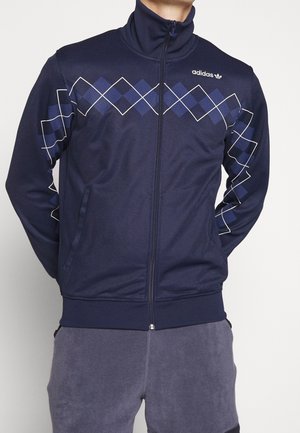 Hombre vestido con una chaqueta de chándal de cremallera de Adidas en azul marino, con un patrón de rombos en azul oscuro y blanco a través del pecho y las mangas.
