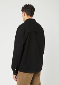 Man die zich van de camera afkeert, gekleed in een zwart overhemd met lange mouwen en knoopmanchetten, en een lichtbruine broek, tegen een effen achtergrond.