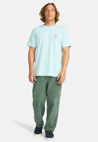 T-shirt in cotone azzurro chiaro con maniche corte e grafica sul petto, abbinato a pantaloni in corduroy verdi e scarpe blu navy. Vestibilità casual e stile rilassato.
