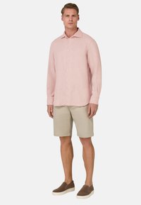Camicia rosa chiaro a maniche lunghe con bottone frontale, pantaloni corti beige e scarpe slip-on marroni. La camicia presenta un colletto standard e una vestibilità rilassata.