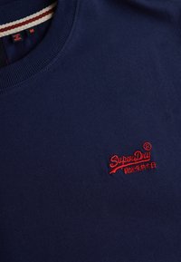 Navy blue crewneck sweater with red embroidered "SuperDry" logo and Japanese text on front, showing collar with size and country tags.