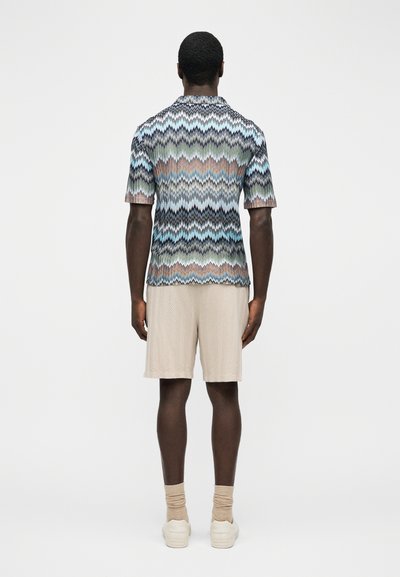 Άνδρας με σκούρο δέρμα που φοράει πολύχρωμη κοντομάνικη μπλούζα με σχέδιο ζιγκ-ζαγκ, μπεζ σορτς, κάλτσες που ταιριάζουν και λευκά αθλητικά παπούτσια, όρθιος με την πλάτη γυρισμένη.