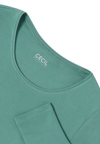 Turquoise kortmouwtop gemaakt van zachte stof, met een ronde halslijn en een pocketdetail aan de voorkant. Label zichtbaar.