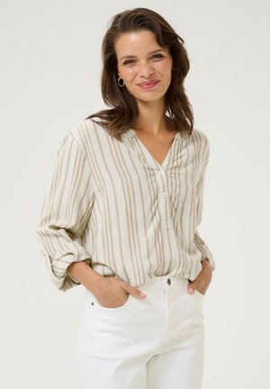Blusa leggera a righe in beige e crema con scollatura a V e tre bottoni, abbinata a pantaloni bianchi. Vestibilità rilassata con maniche arrotolate.