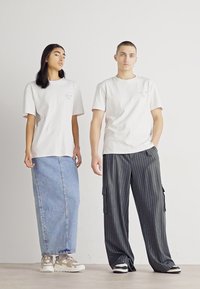 Dos personas están de pie contra una pared blanca vistiendo camisetas blancas; una lleva una larga falda de mezclilla y zapatillas, mientras que la otra lleva pantalones anchos de rayas y zapatillas.