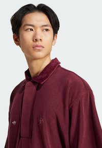 Polo de forro burdeos con un cuello texturizado, que presenta un pequeño logo de Adidas y un detalle bordado en el pecho. Manga corta, corte relajado.