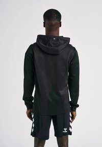 Hoodie nera con un design lungo e una cucitura centrale, abbinata a pantaloni corti neri con motivi a chevron bianchi. Texture liscia, stile atletico moderno.