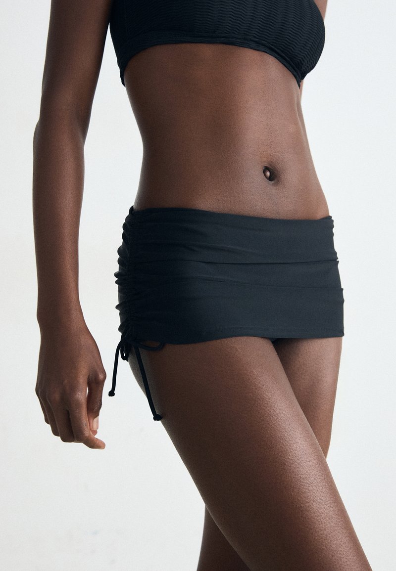 Schwarzer Schwimmrock mit geraffter Textur, verstellbaren Kordeln an den Seiten, glattem Stoff, getragen mit einem passenden Oberteil, das den Bauch zeigt.