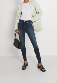 Smala jeans i mörkblå denim, kombinerat med en vit figurnära topp och en ljusgrön oversized kavaj. Svarta klumpiga klacksandaler och en svart handväska.