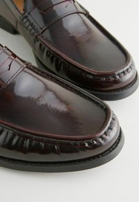Mocasines de cuero burdeos con acabado brillante, con costuras decorativas y forma de puntera redondeada, montados sobre una suela de goma negra.