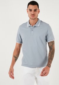 Světle šedé polo tričko vyrobené z hladké tkaniny, s límečkem a bílým lemem a zapínáním na tři knoflíky.