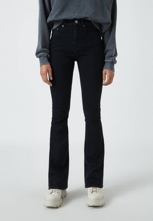 Persoon die donkere, hoog getailleerde flared jeans draagt, een grijze longsleeve trui en witte sneakers met dikke zolen, staand op een lichte achtergrond.