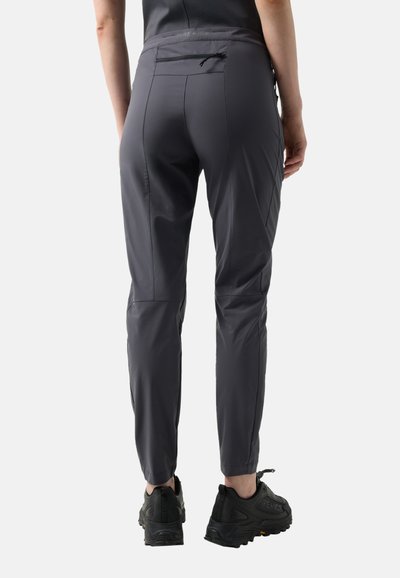 Osoba oblečená v tmavě šedých úzkých kalhotách a černých sportovních botách stojí zády k fotoaparátu na jednobarevném pozadí.