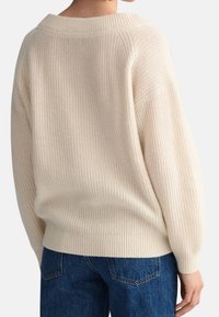 Cremfarbener Strickpullover mit weitem Kragen und rippenartiger Textur, ausgestattet mit überschnittenen Schultern und engen Manschetten. Kombiniert mit dunkelblauen Jeans.