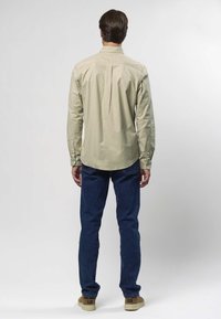 Hellbeiges Langarmhemd mit abgerundetem Saum und Rückenteil, kombiniert mit dunkelblauen Jeans und hellbraunen Sneakers. Ansicht von hinten.