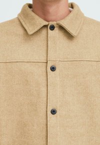 Beige wool-blend jacket with a textured finish, featuring a collar, front button closure, and two black buttons on the placket.