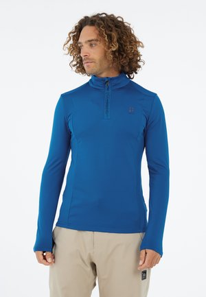 Mann mit lockigem Haar, der ein blaues langärmliges Zipshirt und beige Hosen trägt, schaut zur Seite vor einem weißen Hintergrund.