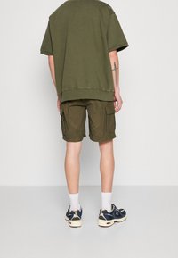 Camiseta oversized de color verde oliva combinada con pantalones cortos de cargo en verde oliva con bolsillos laterales. Calcetines blancos y zapatillas con detalles en azul.