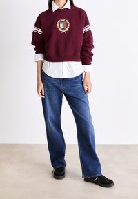 Sudadera burdeos con rayas blancas, con un parche de logo. Llevada sobre una camisa blanca de cuello y combinada con jeans azules y zapatillas negras.