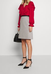 Punainen röyhelöblusa nappikiinnityksellä, harmaa kuvioitu hame, mustat nilkkaketjulliset kengät ja pieni musta käsilaukku.