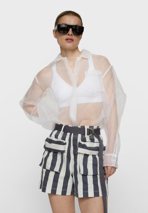 Mujer con gafas de sol negras extragrandes, camisa blanca transparente con botones, sujetador blanco y pantalones cortos de rayas azul marino y blanco de talle alto con grandes bolsillos.