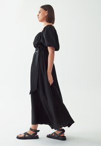 Black, off-shoulder dress with a textured fabric, wide sleeves, and a belted waist. Worn with black strappy sandals.