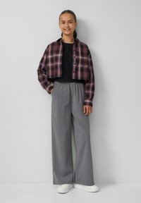 Geknipte geruite blouse in paars en zwart over een zwarte top, gecombineerd met grijze wijde broek en witte sneakers, handen in de zakken.