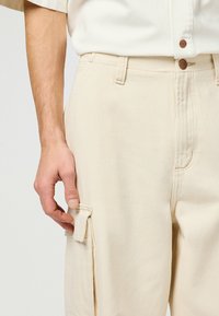 Beige katoenen cargobroek met een zijzak, knoopsluiting en ri loops. De stof heeft een gladde textuur en zichtbare stikseldetails.