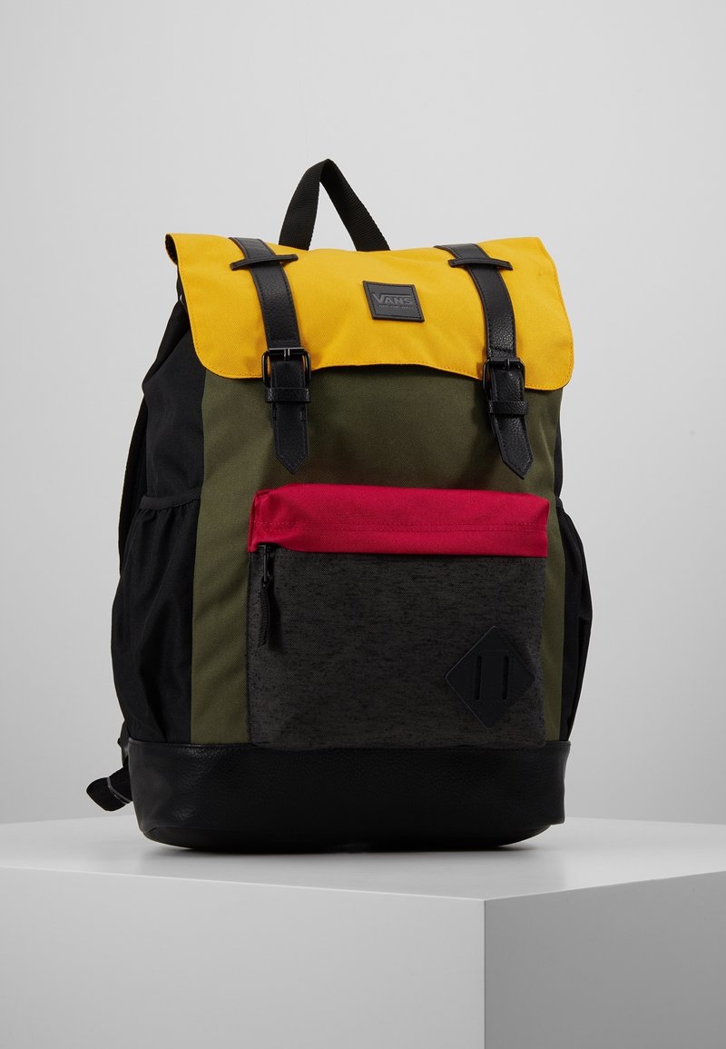 Sac à dos avec rabat supérieur jaune, bretelles noires, corps vert, poche avant rouge et fond noir, présenté sur un socle blanc sur fond gris.