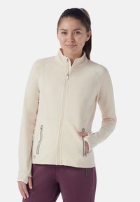 Giacca zip in beige chiaro con colletto alto, due tasche laterali con zip e design aderente. Tessuto morbido e elasticizzato con cuciture piatte.