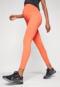 Neon oransje leggings med slim passform og høy midje, med subtile sømdetaljer, kombinert med svarte joggesko med hvite aksenter.