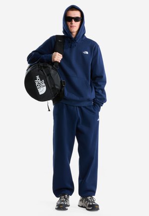 Mann trägt eine marineblaue The North Face Kapuzenjacke und Hose, schwarze Sonnenbrille, graue Sneakers, trägt eine schwarze runde The North Face Tasche auf der Schulter.