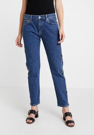 Femme portant un jean bleu taille haute avec un ruban logo Calvin Klein le long des coutures latérales et des sandales à talons noires ornées de clous argentés.